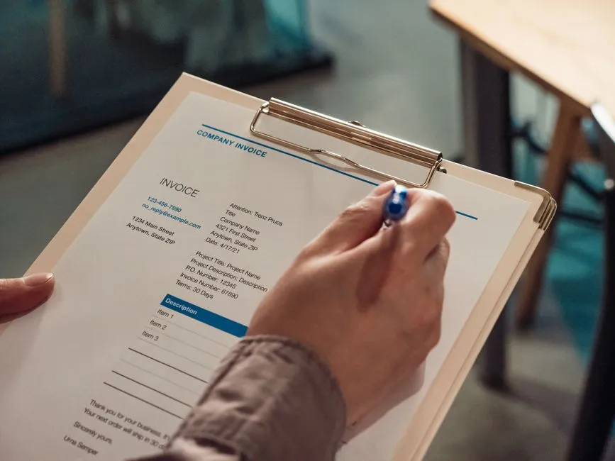 Main tenant un stylo au-dessus d'une facture d'entreprise imprimée sur un porte-bloc, avec coordonnées, numéro de facture et lignes de prestations