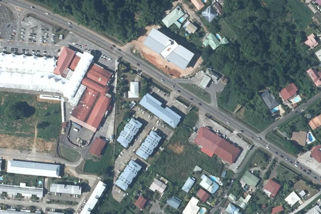 Vue aérienne verticale de la route de Montjoly, à Rémire-Montjoly : quatre petits immeubles d'habitation alignés avec leurs rangées de stationnement, entre un centre commercial et un tissu de villas