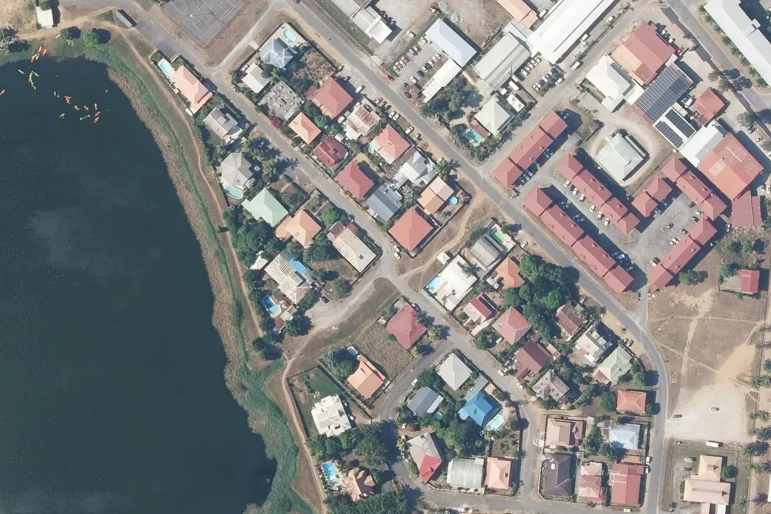 Vue aérienne verticale du quartier Goupi à Kourou : un ensemble de logements collectifs à toitures rouges avec ses parkings, en bordure d'un lac, face à un lotissement de villas