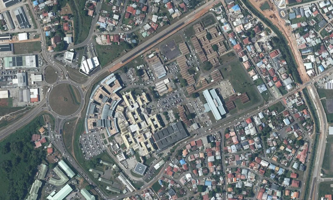 Vue aérienne verticale du centre hospitalier de Cayenne, avenue des Flamboyants : le bâtiment en arc de cercle à verrières sur la gauche, les blocs d'hospitalisation crème alignés au centre, les nappes de stationnement du personnel à droite, et le rond-point de la rocade dans l'angle gauche