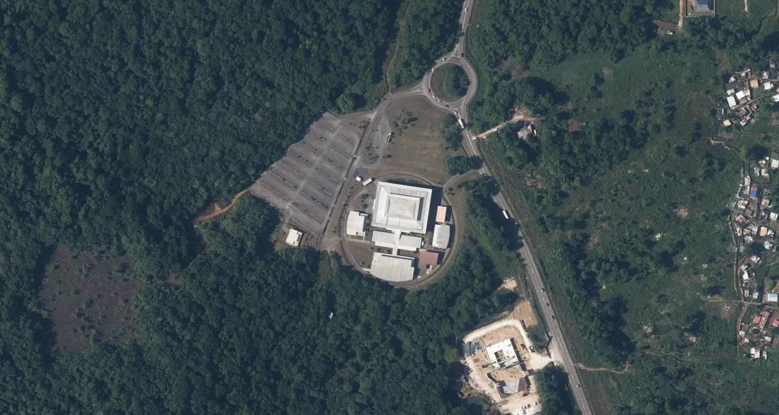 Vue aérienne du Palais régional omnisports Georges-Théolade à Matoury : la halle carrée à toiture pyramidale entourée de sa voie circulaire, les rangées de stationnements couverts sur la gauche, la RN 2 et son rond-point sur la droite, la forêt tout autour