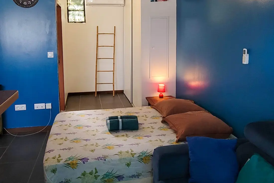 Studio du Sweet Dreams : mur bleu, lit à parure fleurie, serviettes pliées, échelle en bambou et climatiseur avec télécommande