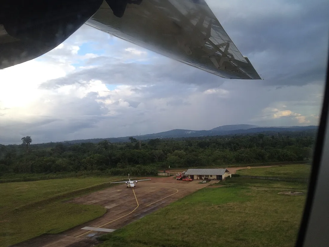 Vue depuis le hublot d'un avion en approche de l'aérodrome de Maripasoula : sous l'aile, une piste unique en dur bordée d'herbe, un bimoteur blanc stationné sur l'aire de trafic, un bâtiment bas à toiture de tôle et quelques véhicules, puis la forêt dense et les collines à l'horizon