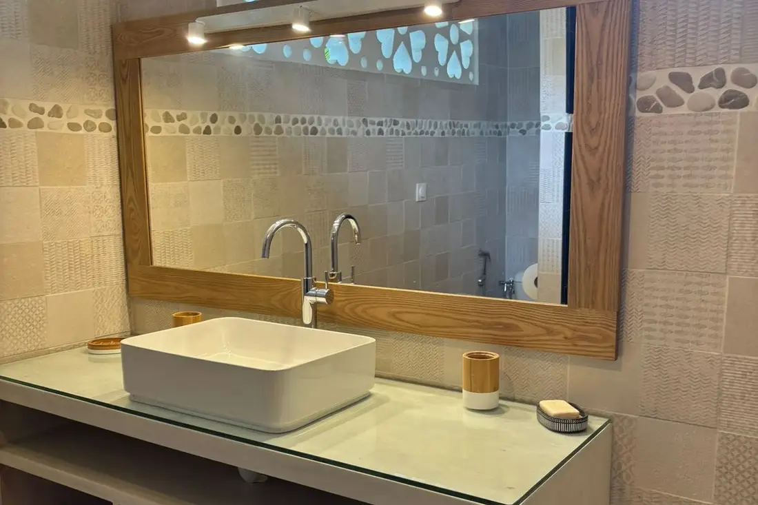 Salle de bain de la maison Tropikana : vasque blanche posée sur plan vitré, grand miroir à cadre en bois, frise de galets