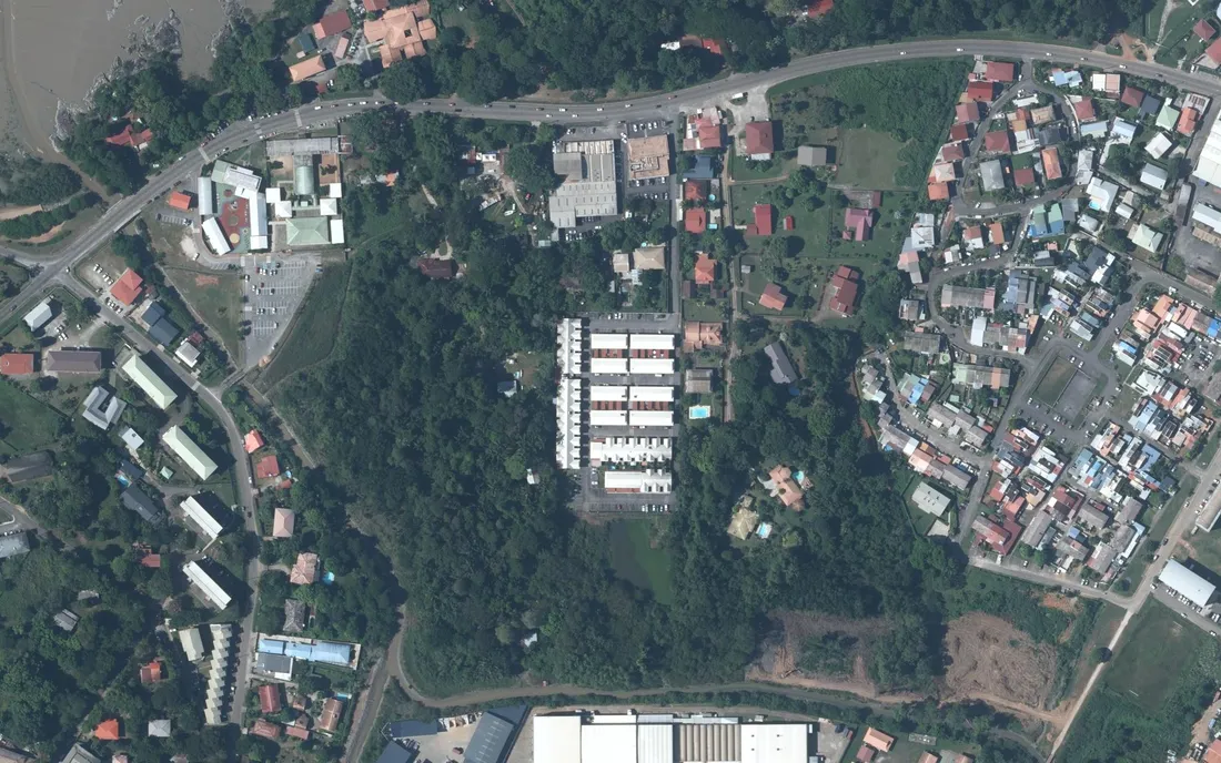 Vue aérienne verticale du secteur du 494 route de Montabo, à Cayenne : au centre, l'ensemble de maisons mitoyennes du Clos d'Alpinias enserré de forêt ; en haut, la route de Montabo à quatre voies ; à droite, le quartier pavillonnaire dense ; en haut à gauche, la vasière de l'estuaire