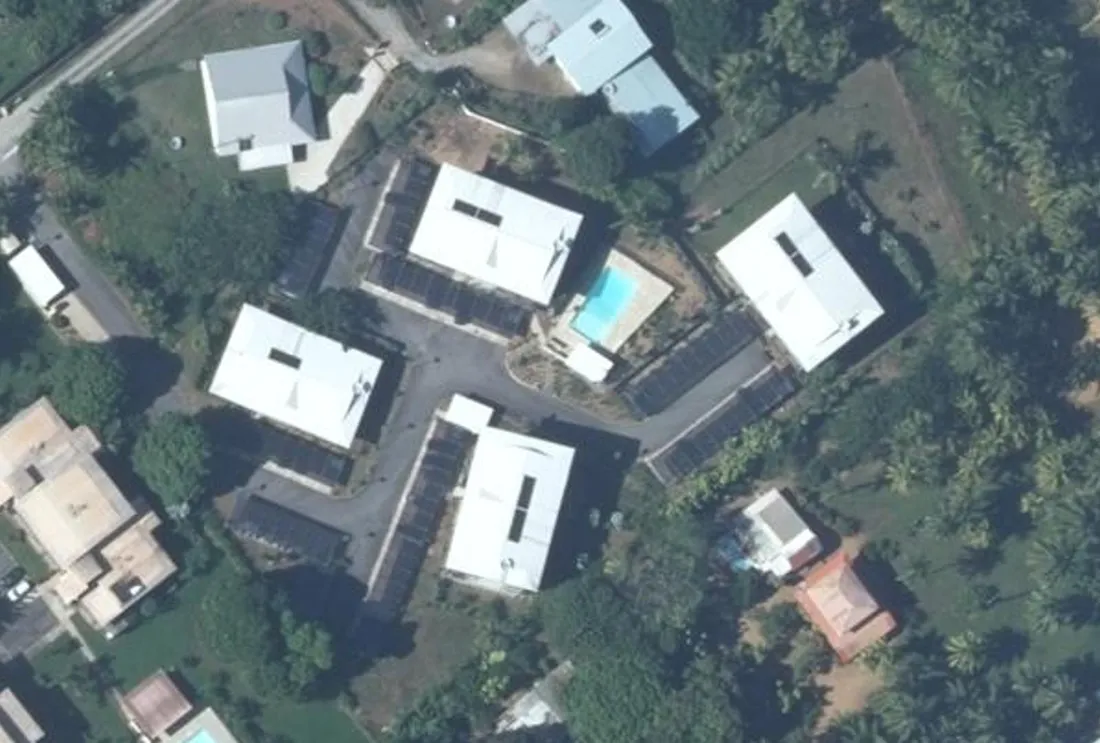 Vue aérienne verticale de la résidence Coconut Plage, 6 rue Canne à Sucre à Cayenne : quatre bâtiments à toiture blanche autour d'une allée de desserte, longues rangées de stationnements couverts et piscine
