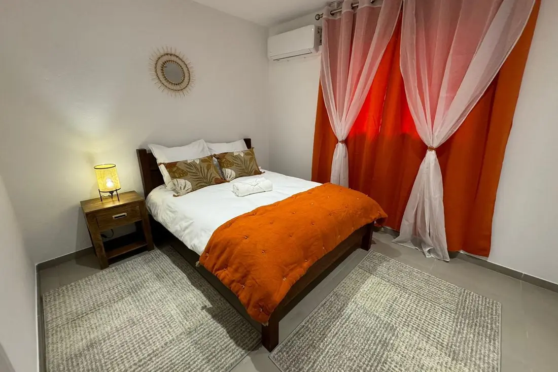 Chambre d’un appartement de la Résidence Francis : climatiseur mural, lit à couvre-lit orange, rideaux orangés et lampe en rotin