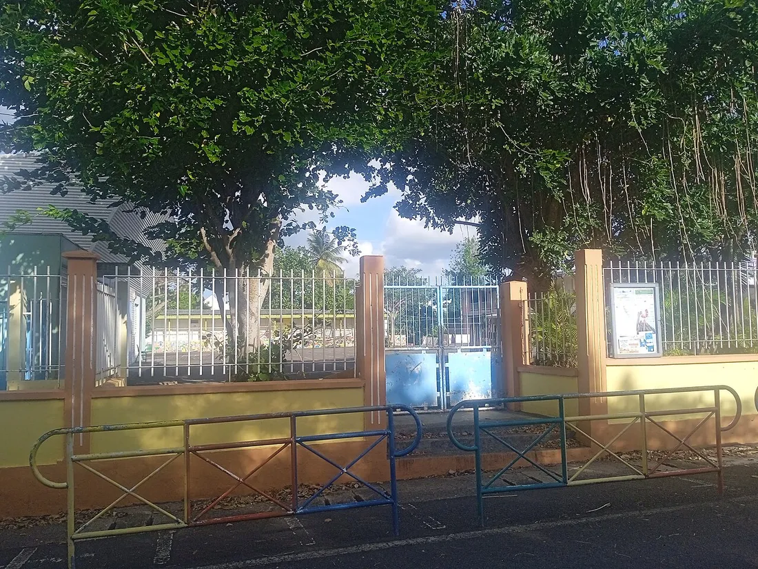 Entrée grillagée de l'école du Bourg de Sainte-Rose en Guadeloupe, avec barrières colorées et panneau d'affichage sous les arbres