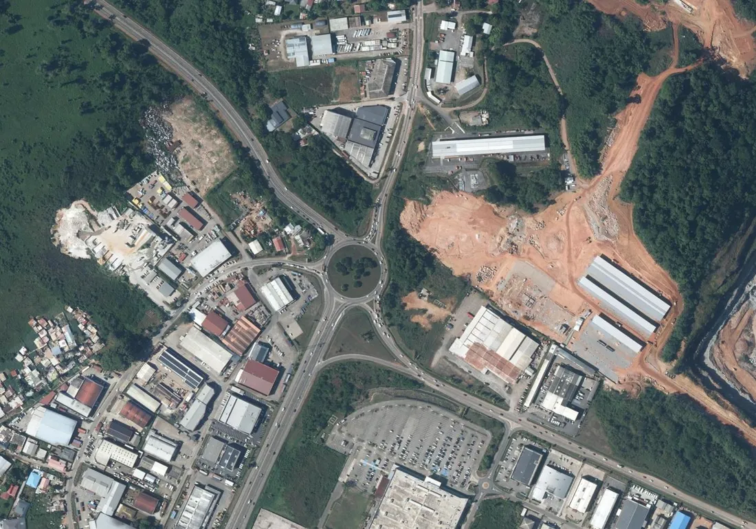 Vue aérienne verticale du giratoire des Maringouins, à l'ouest de Cayenne : le grand rond-point planté d'arbres au croisement de la route nationale, la zone d'activités et ses hangars à gauche, l'ancienne carrière aux terrains rouges et les bâtiments neufs à droite, un vaste parking en bas