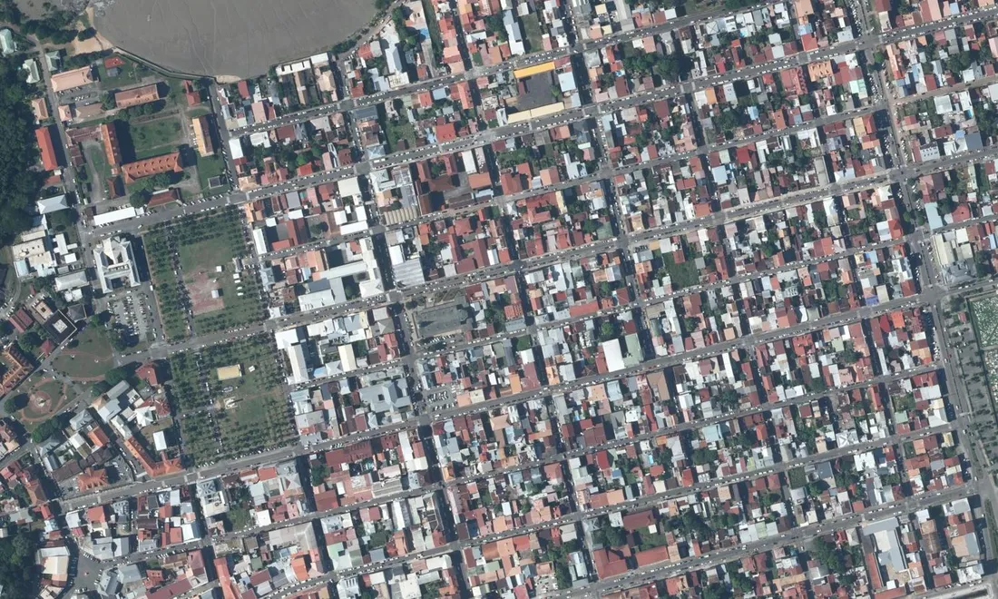 Vue aérienne verticale du centre historique de Cayenne : la place des Palmistes, grand rectangle vert planté de rangées de palmiers en alignement, entourée du damier serré des rues et des toitures rouges et grises ; la vasière de l'estuaire en haut à gauche, le cimetière en bordure droite