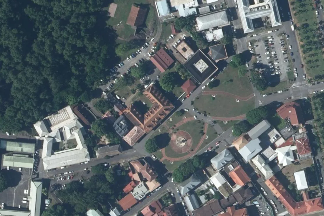 Orthophotographie verticale de la place Léopold-Héder à Cayenne : le bâtiment de la préfecture de la région Guyane, sa cour et ses toits de tôle, la place plantée avec son kiosque circulaire, et le versant boisé du Fort Cépérou