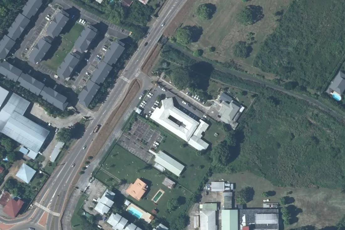 Orthophotographie verticale de la rocade de Zéphir (RD18) à Cayenne : la deux-voies, un ensemble de bâtiments administratifs blancs à cour intérieure avec son parking, et des résidences aux toits gris sur le côté opposé