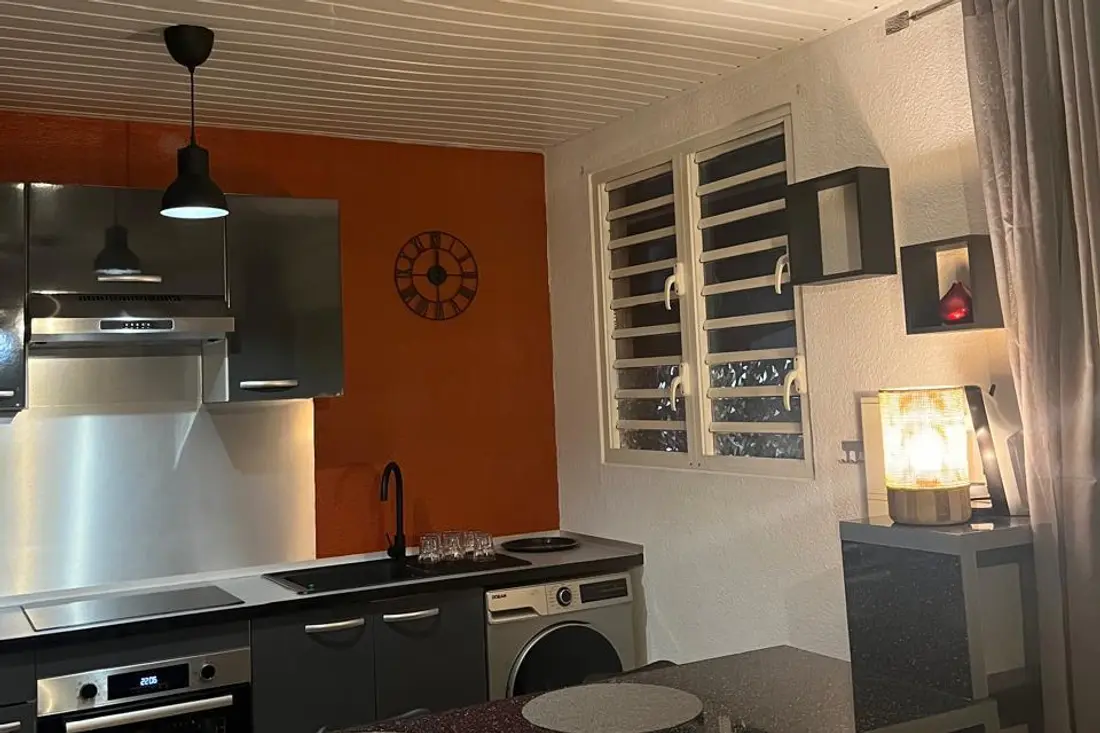 Cuisine d’un appartement Cayenne Chic le soir : mur orange, hotte inox, lave-linge et lampe d’appoint allumée près de la fenêtre à jalousies