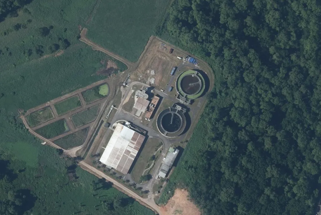 Vue aérienne verticale d'une station d'épuration isolée en lisière de forêt à Cayenne : deux bassins circulaires en béton, un grand hangar couvert de lits de séchage des boues et des bassins rectangulaires en terre