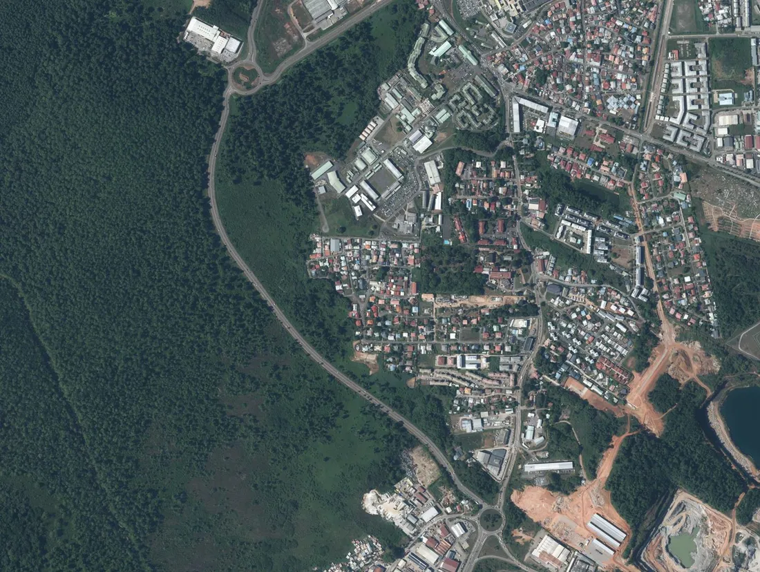 Vue aérienne verticale de la RN1 entre le giratoire Leblond et le giratoire des Maringouins à Cayenne : une chaussée unique longe le marais Leblond, la ville s'étendant sur toute la droite