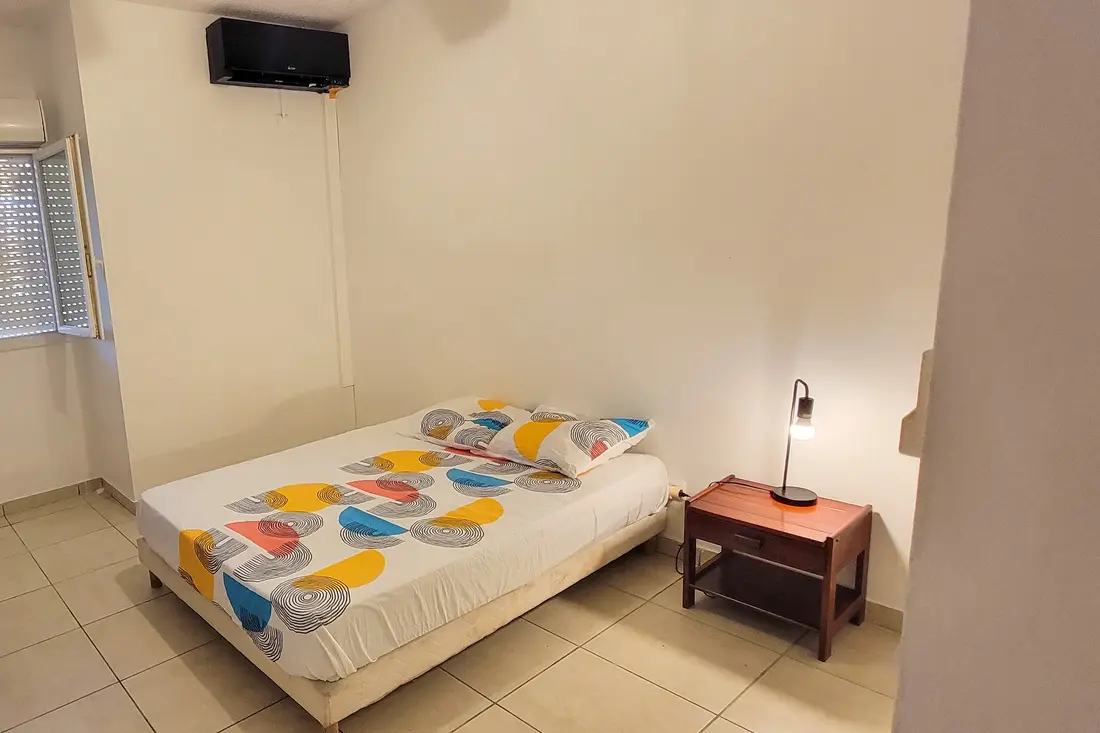 Chambre de la Villa Mahury : climatiseur au mur, lit double à motifs colorés et table de chevet en bois