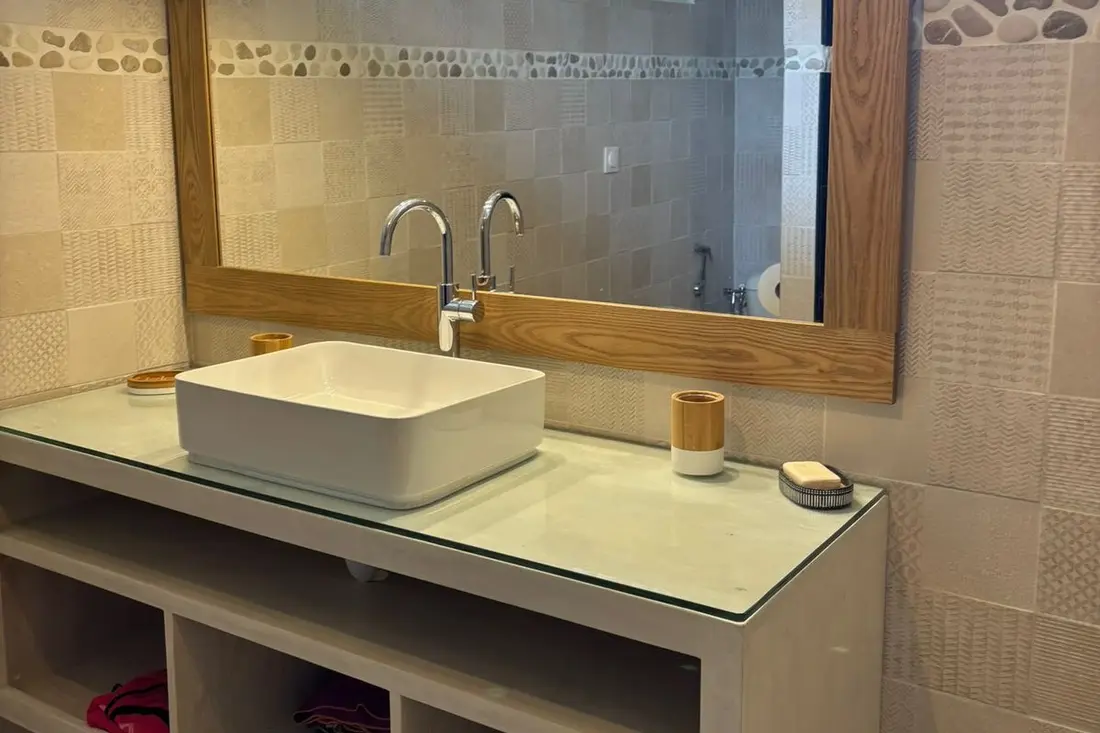 Salle de bain de la Tropikana : vasque blanche sur plan vitré, grand miroir à cadre bois et frise de galets
