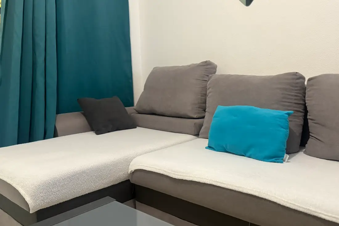 Canapé d’angle du Diamant Cayennais : assises claires, coussins gris et bleu canard devant le rideau